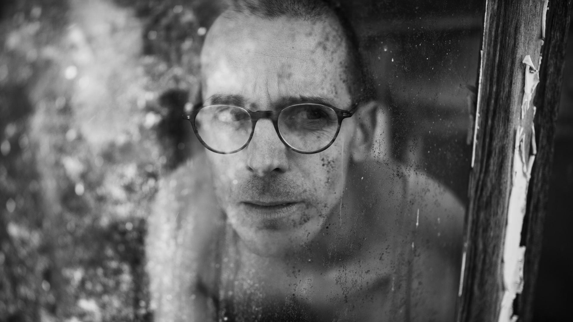 Ein Mann mt Brille und nacktem Oberkörper hinter eine Fensterscheibe