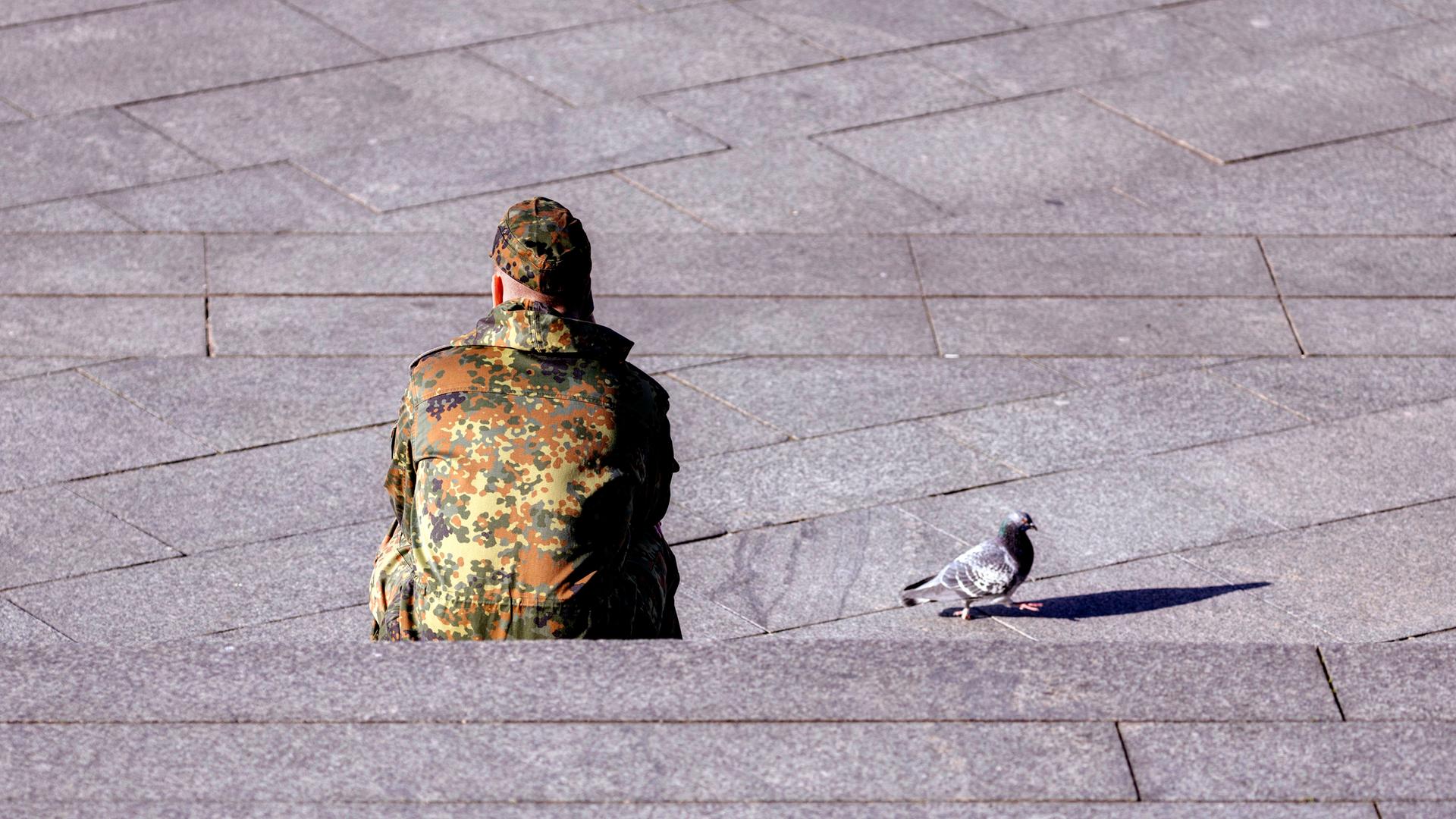 Ein Bundeswehr-Soldat in Tarnkleidung sitzt mit dem Rücken auf einer Treppe. Daneben ist eine Taube zu sehen.