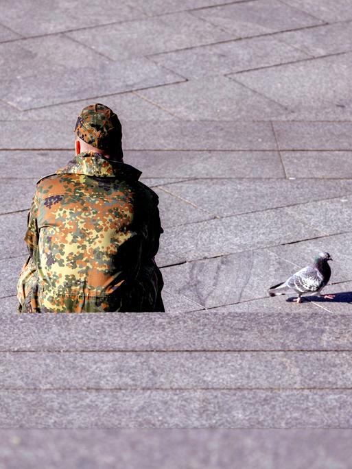 Ein Bundeswehr-Soldat in Tarnkleidung sitzt mit dem Rücken auf einer Treppe. Daneben ist eine Taube zu sehen.