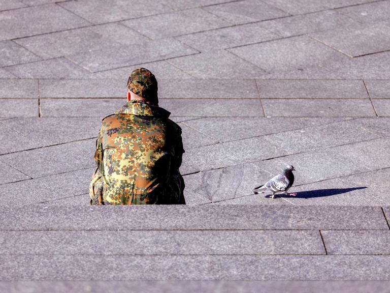 Ein Bundeswehr-Soldat in Tarnkleidung sitzt mit dem Rücken auf einer Treppe. Daneben ist eine Taube zu sehen.