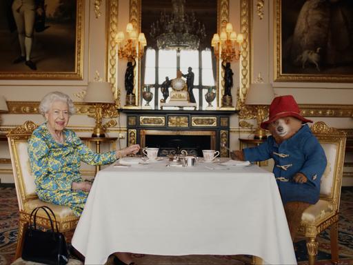 Queen Elizabeth II. und ein Darsteller der Kinderbuchfigur Paddington sitzen im Buckinghampalast an einem Tisch.
