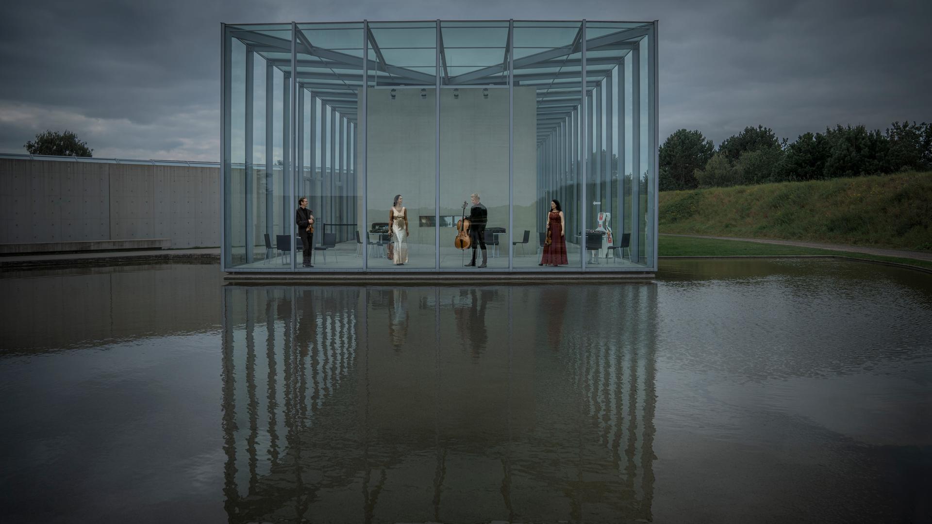 Musikerinnen und Musiker eines Streichquartetts stehen in einem Raum aus Glas, der umgeben von Wasser ist. Der Himmel darüber ist bewölkt.