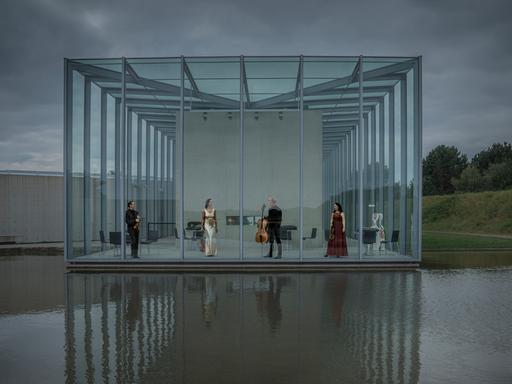 Die vier Musiker des Quartetts stehen in einer Art Glashalle, die zum Wasser hin komplett offen ist und sich im Wasser spiegelt.