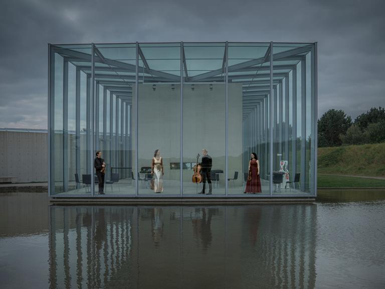Die vier Musiker des Quartetts stehen in einer Art Glashalle, die zum Wasser hin komplett offen ist und sich im Wasser spiegelt.