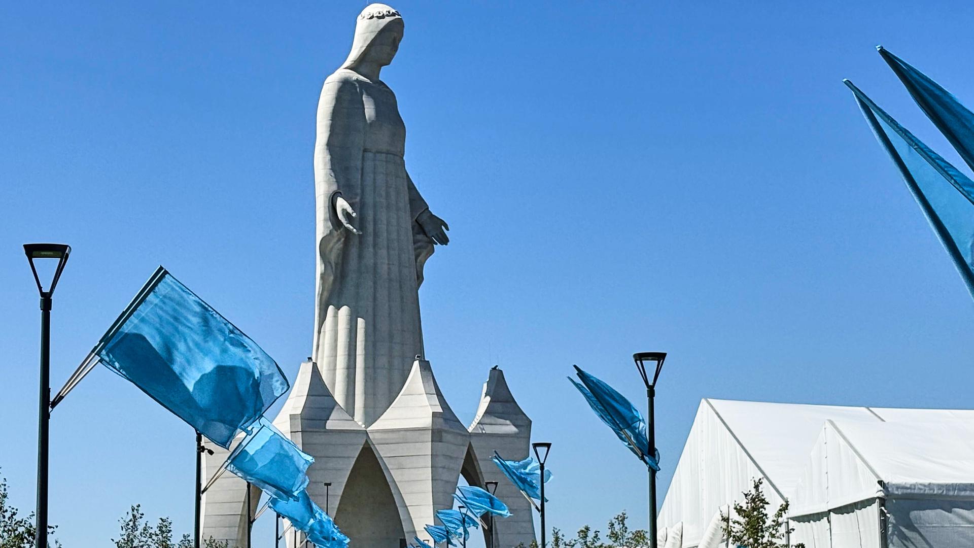 Auf dem Weg zu der riesigen Statue stehen unzählige blaue Fahnen. Darüber ein stahlblauer wolkenloser Himmel. 