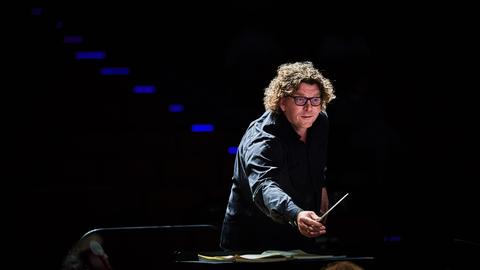 Ein Mann in schwarzem Hemd steht mit Dirigierstab am Pult, fotografiert aus Orchesterperspektive.