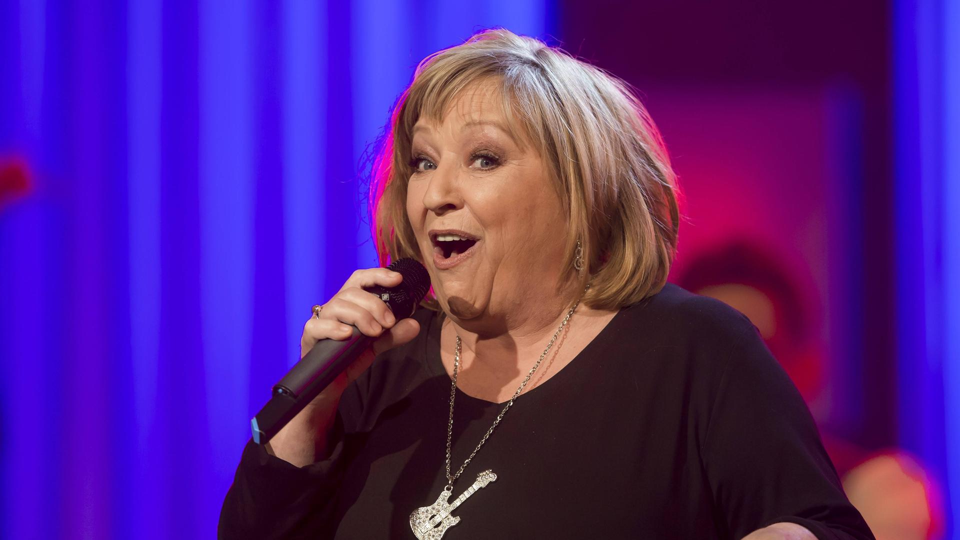 Die Sängerin Angelika Mann steht vor einem blauen Hintergrund auf einer Bühne und singt in ein Mikrofon. An ihrer Halskette baumelt eine Schmuck in Gitarrenform.