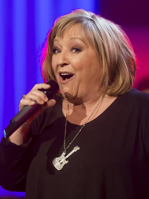 Die Sängerin Angelika Mann steht vor einem blauen Hintergrund auf einer Bühne und singt in ein Mikrofon. An ihrer Halskette baumelt eine Schmuck in Gitarrenform.