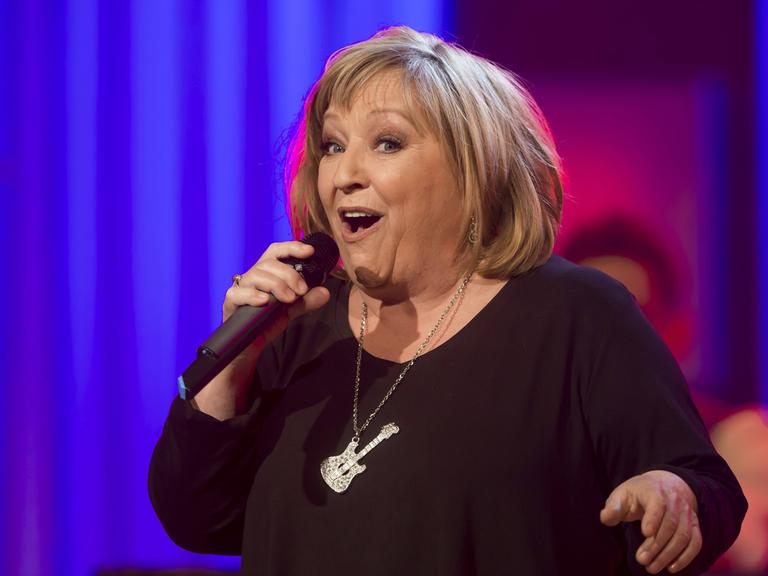 Die Sängerin Angelika Mann steht vor einem blauen Hintergrund auf einer Bühne und singt in ein Mikrofon. An ihrer Halskette baumelt eine Schmuck in Gitarrenform.