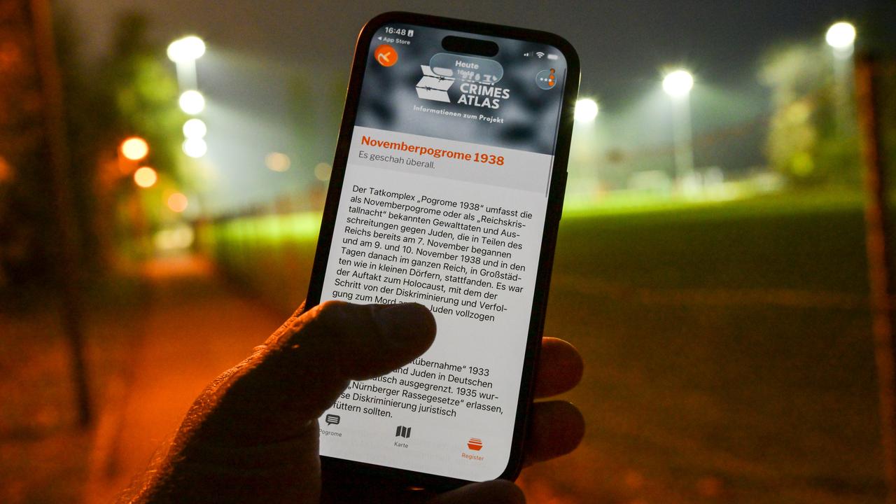 Auf einem Smartphone ist die App "NaziCrimesAtlas" geöffnet. Ein Mensch hält das Handy an einer Straße vor sich. Es ist dunkel, im Hintergrund ein beleuchteter Sportplatz.