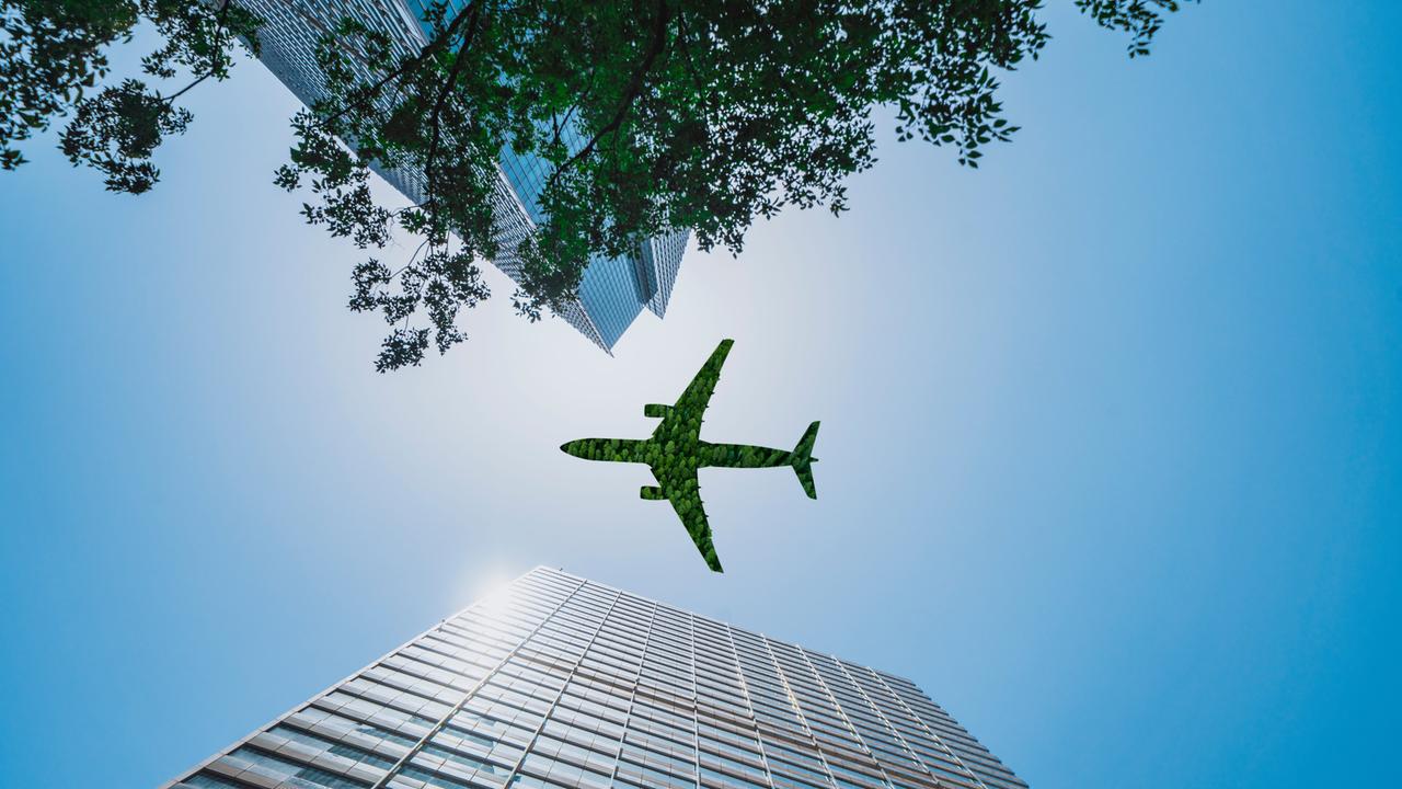 Aufnahme eines grünen Flugzeugs, das über ein Bürogebäude fliegt, aus der Froschperspektive, das Flugzeug sieht aus, als wäre es aus Baumgrün gefertigt