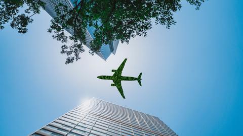 Aufnahme eines grünen Flugzeugs, das über ein Bürogebäude fliegt, aus der Froschperspektive, das Flugzeug sieht aus, als wäre es aus Baumgrün gefertigt
