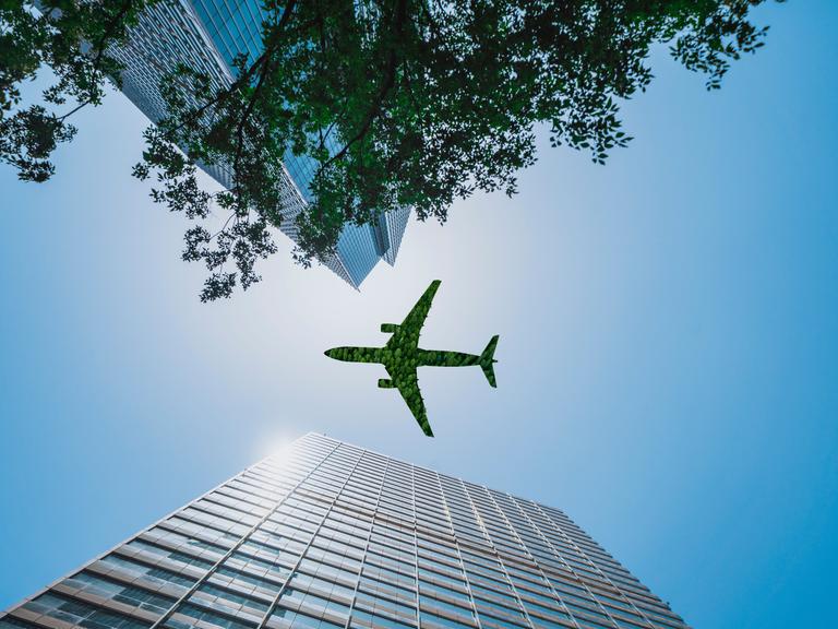 Aufnahme eines grünen Flugzeugs, das über ein Bürogebäude fliegt, aus der Froschperspektive, das Flugzeug sieht aus, als wäre es aus Baumgrün gefertigt