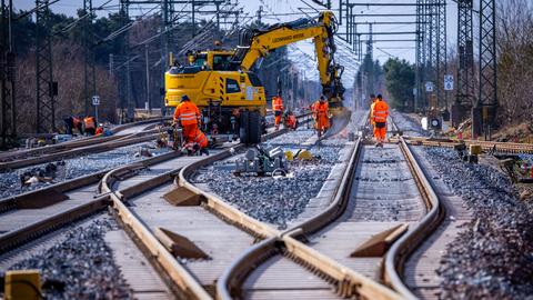 Bauarbeiter montieren mithilfe eines gelben Baggers Gleisanlagen.