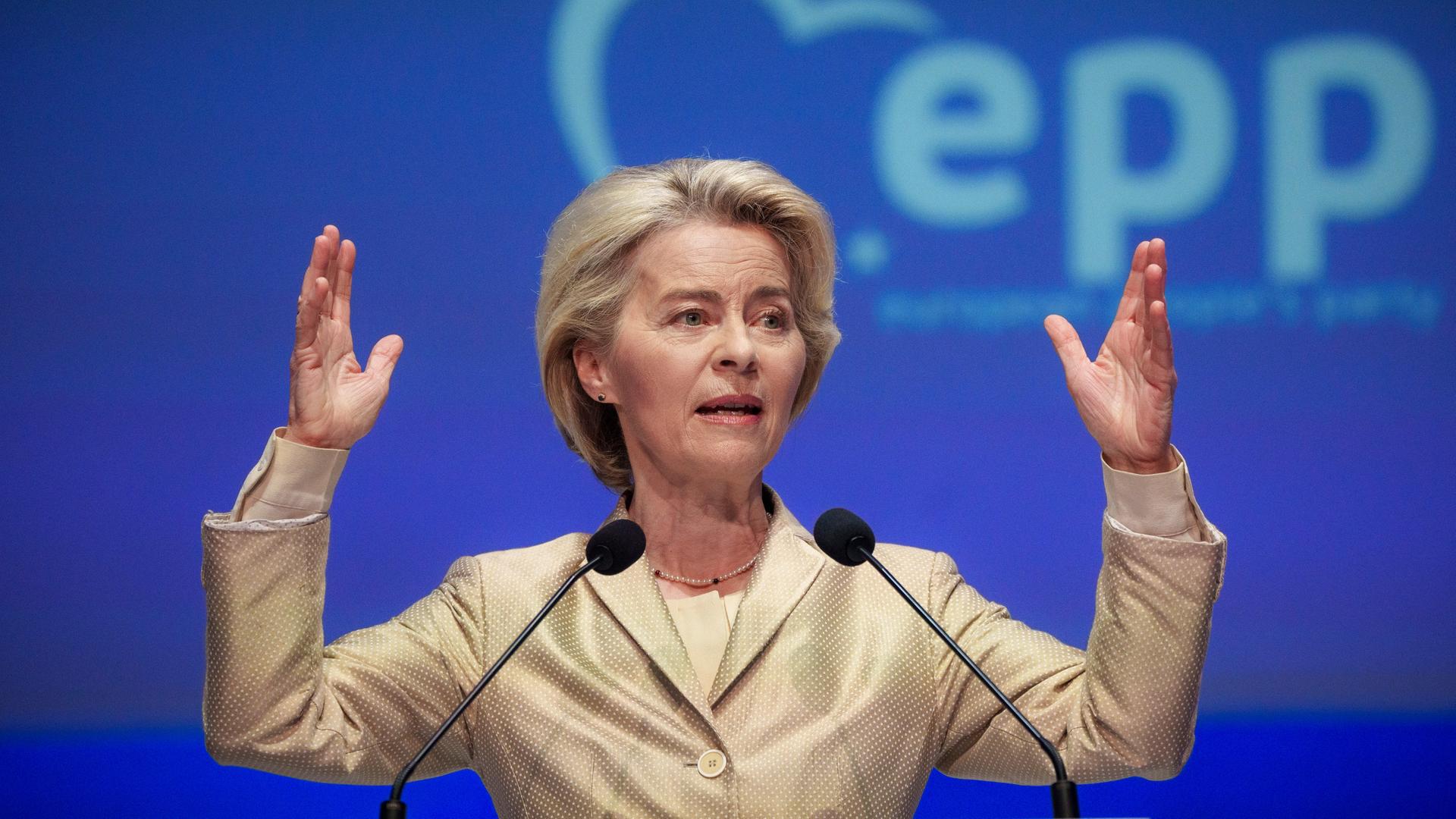 Ursula von der Leyen steht mit erhobenen Händen vor zwei Mikrofonen.