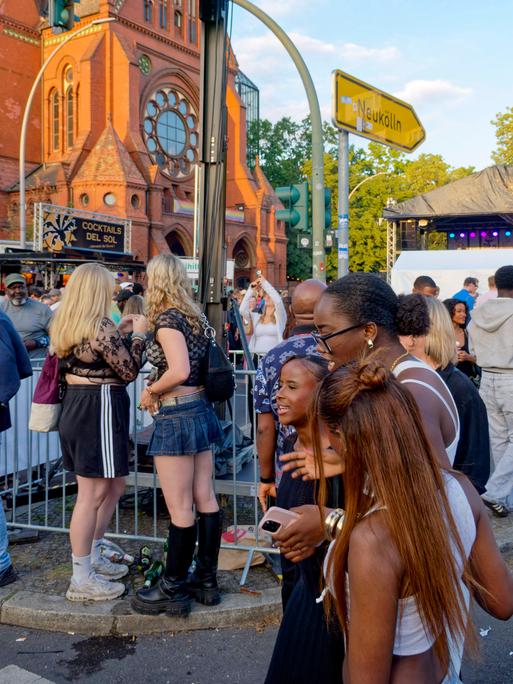 Menschen feiern zusammen auf der Straße. Im Hintergrund ist eine Kirche zu sehen und ein Hinweisschild "Neukölln".