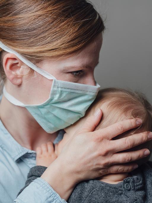 Eine Mutter mit medizinischer Maske trägt ihr krankes Kleinkind auf dem Arm.