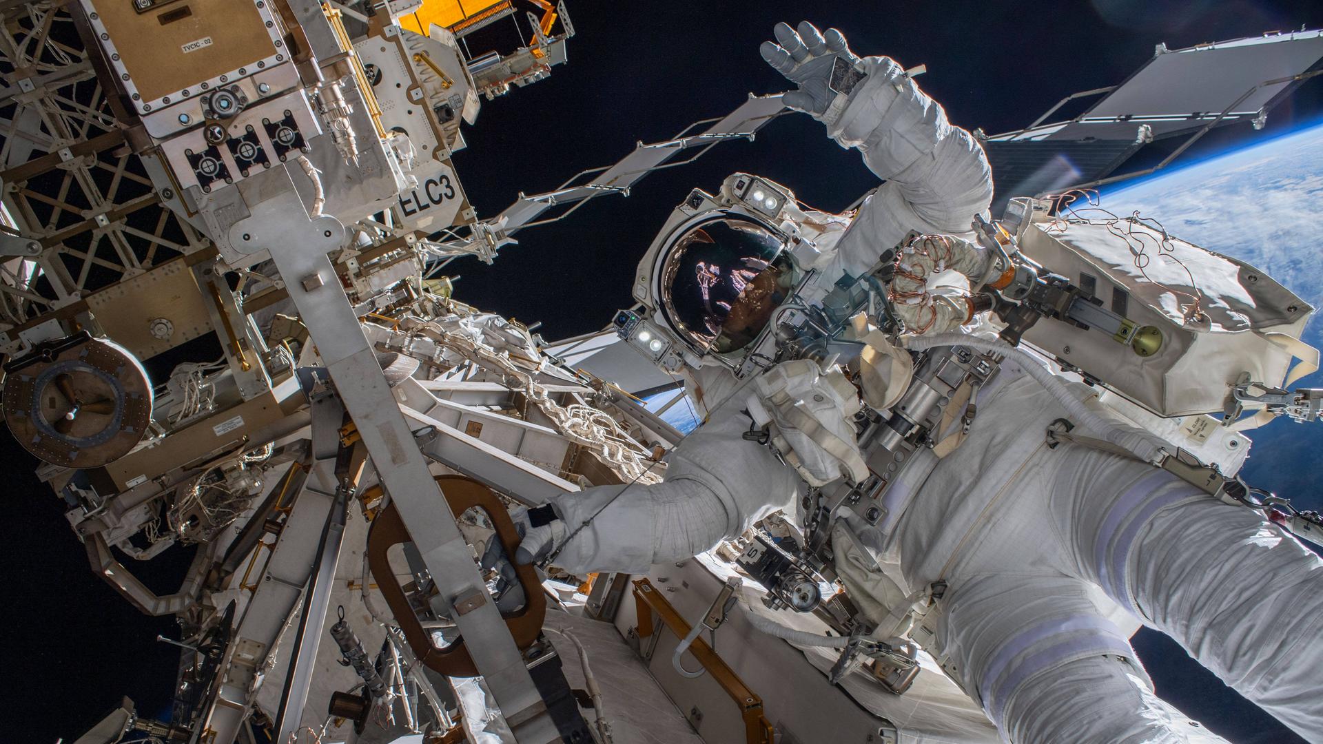 Der Astronaut Matthias Maurer arbeitet außerhalb der Raumstation ISS und winkt in die Kamera. Im Hintergrund ist die Erde zu sehen