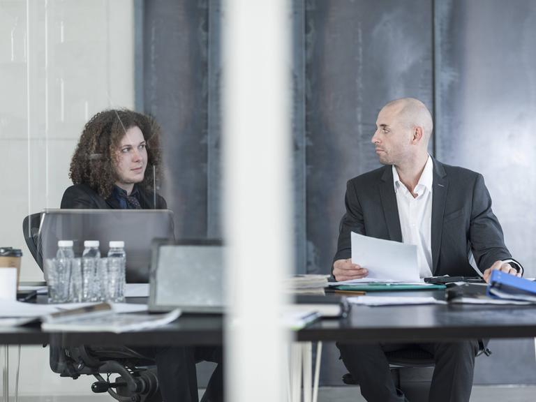 Zwei Personen im Business-Dress - eine jung, die andere etwas älter - sitzen an Schreibtischen und schauen sich skeptisch an.