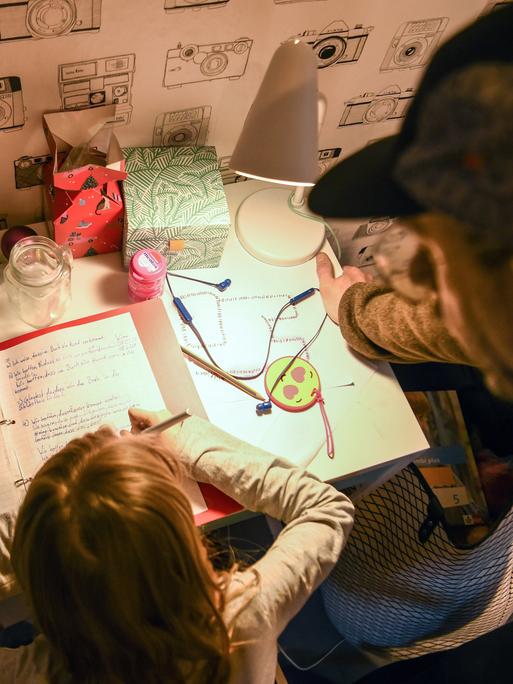 Ein Mädchen sitzt zu Hause an ihrem Schreibtisch und macht Aufgaben für die Schule. Ihr Vater steht daneben und sieht ihr zu.
