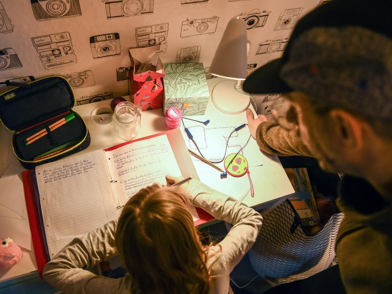 Ein Mädchen sitzt zu Hause an ihrem Schreibtisch und macht Aufgaben für die Schule. Ihr Vater steht daneben und sieht ihr zu.