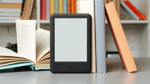 Ein eReader lehnt an einem Regal voller Bücher, daneben ein aufgeschlagenes Buch.