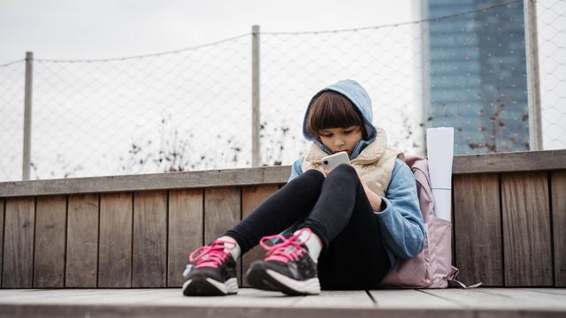 Ein junges Mädchen sitzt allein auf einer Mauer. Es schaut in ein Smartphone.