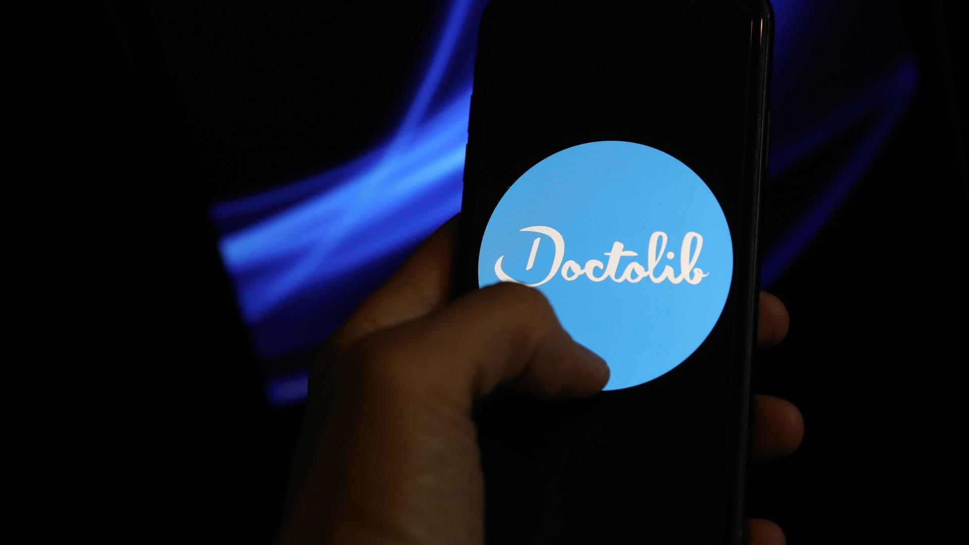 Eine Hand hält ein Smartphone, auf dem das Logo von Doctolib zu sehen ist. 