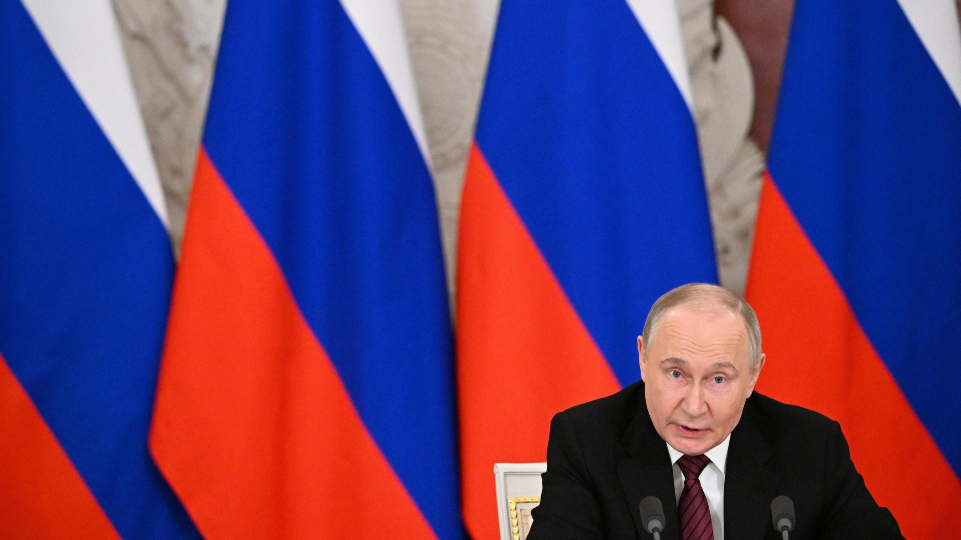 Russlands Präsident Wladimir Putin sitzt vor mehreren russischen Flaggen und spricht in ein Mikrofon.
