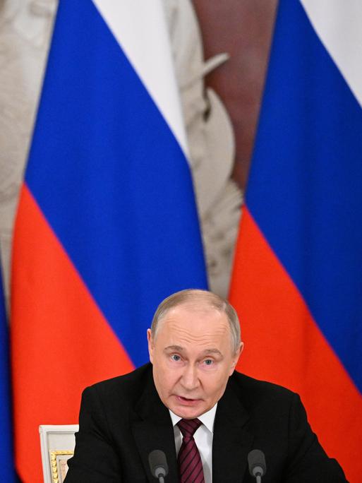 Russlands Präsident Wladimir Putin sitzt vor mehreren russischen Flaggen und spricht in ein Mikrofon.
