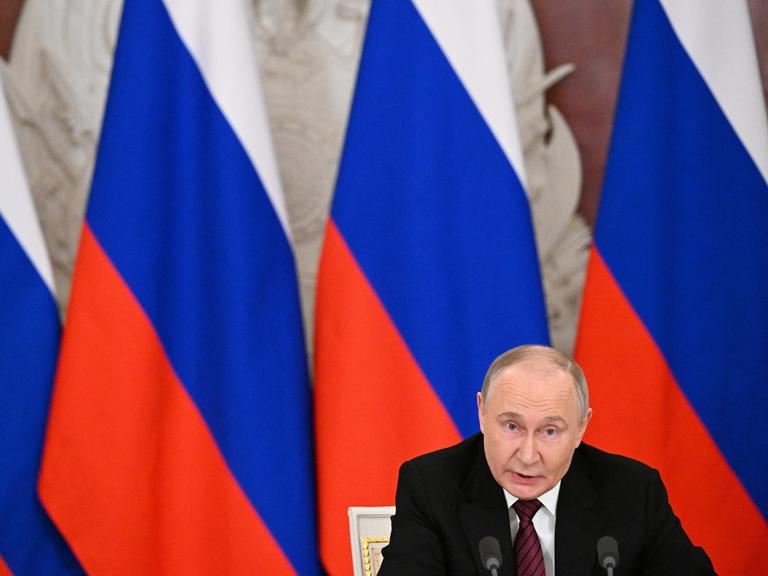 Russlands Präsident Wladimir Putin sitzt vor mehreren russischen Flaggen und spricht in ein Mikrofon.