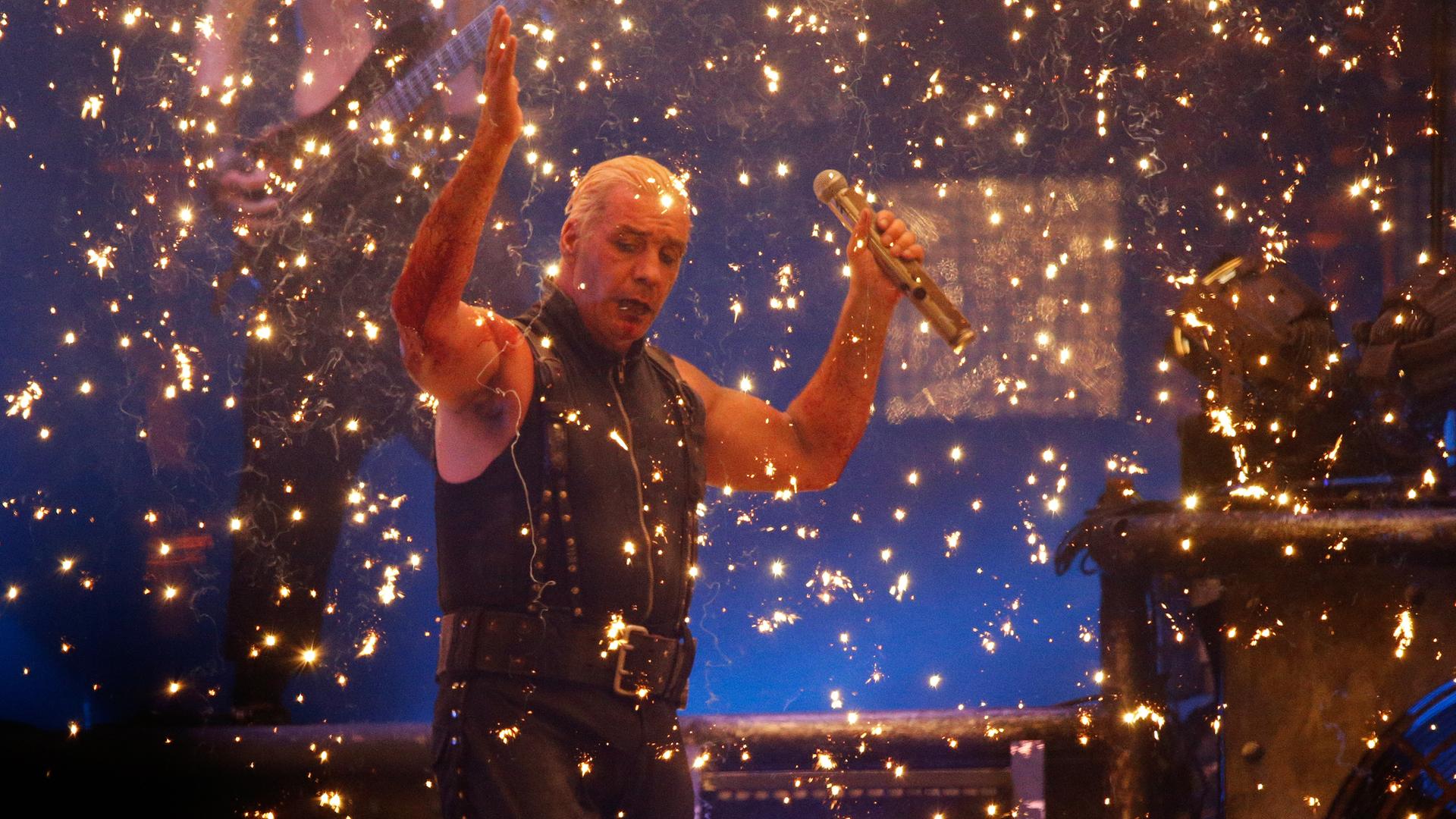 Till Lindemann steht mit erhobenen Händen auf einer Bühne, von goldenem Konfettiregen umgeben