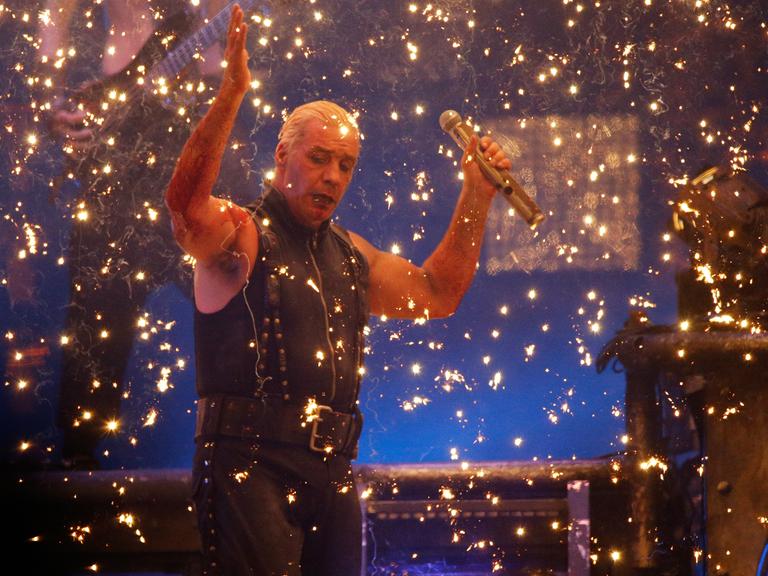 Till Lindemann steht mit erhobenen Händen auf einer Bühne, von goldenem Konfettiregen umgeben
