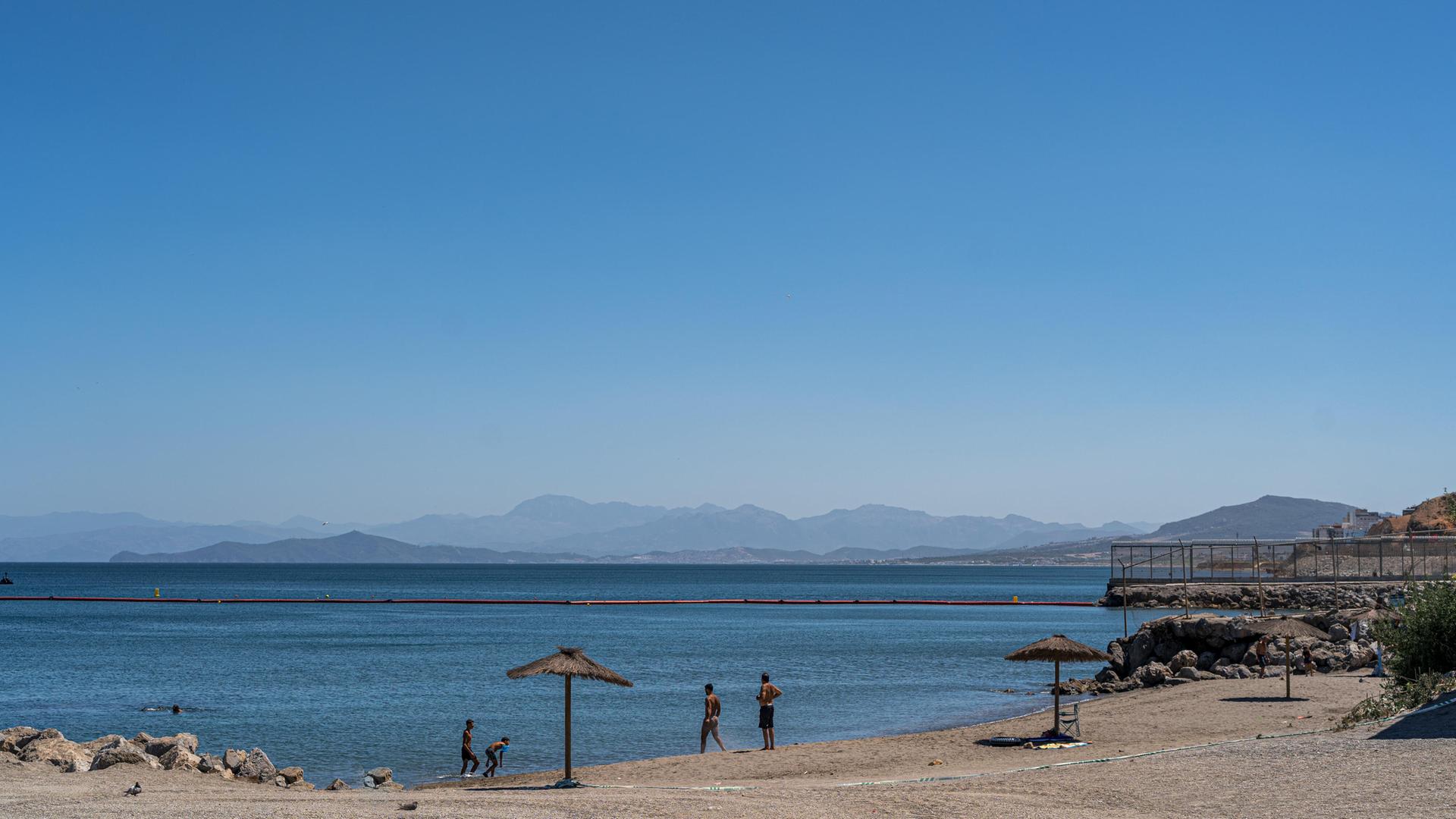 Am Strand von Ceuta baden ein paar Menschen, das Meer ist ruhig und wird von einer Grenzbarriere durchzogen, die sich am Festland mit einem Zaun fortsetzt. Am Strand von Ceuta baden ein paar Menschen, das Meer ist ruhig und wird von einer Grenzbarriere durchzogen, die sich am Festland mit einem Zaun fortsetzt.