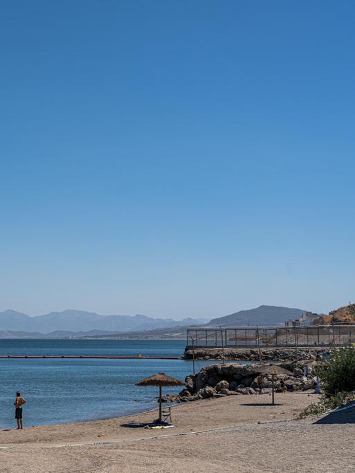 Am Strand von Ceuta baden ein paar Menschen, das Meer ist ruhig und wird von einer Grenzbarriere durchzogen, die  sich am Festland mit einem Zaun fortsetzt.