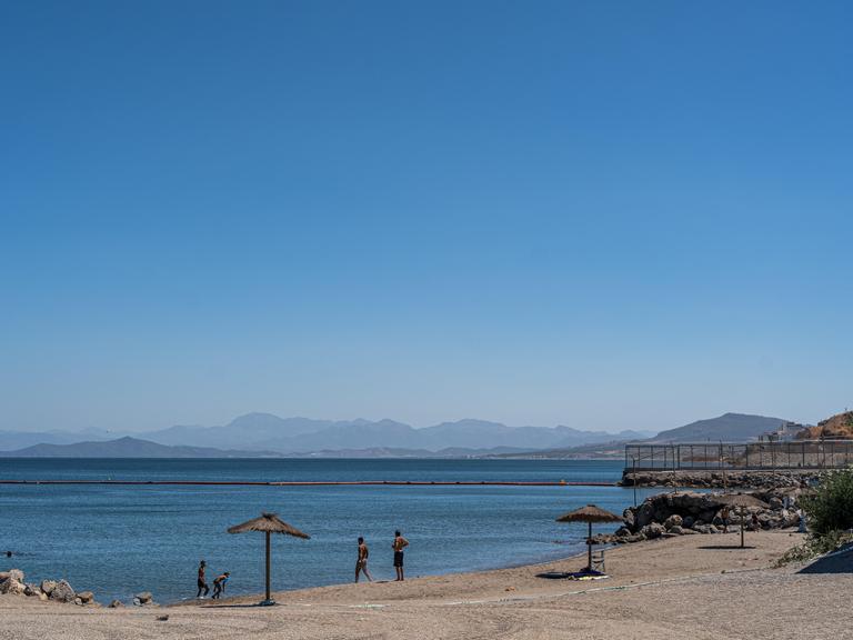 Am Strand von Ceuta baden ein paar Menschen, das Meer ist ruhig und wird von einer Grenzbarriere durchzogen, die  sich am Festland mit einem Zaun fortsetzt.