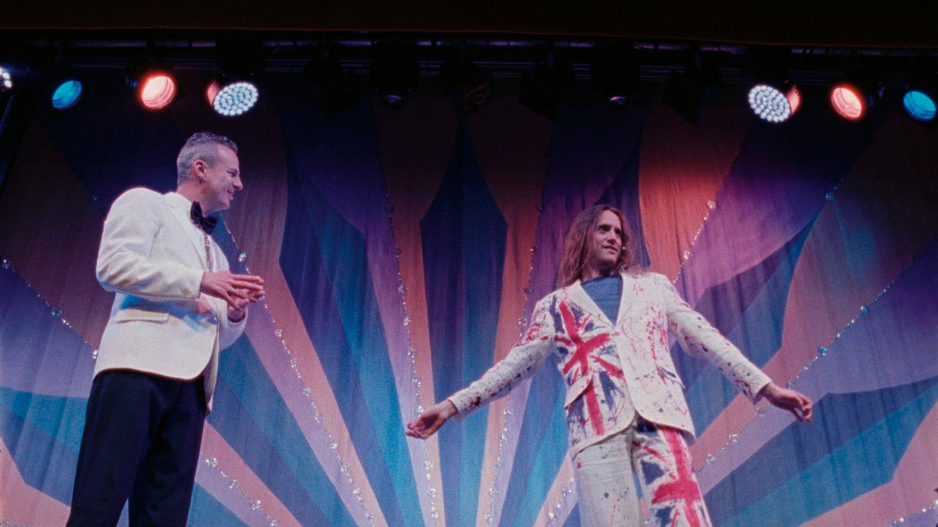 Aus der Doku "Seaside Special": Zwei Personen stehen auf einer Bühne. Eine Person trägt ein Glitzerkostüm mit der britischen Flagge darauf. 