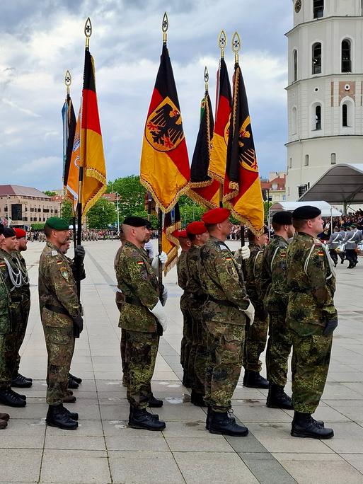 Deutsche Soldaten stehen in Uniformen auf einem öffentlichen Platz in Litauen, einige halten die deutsche Flagge in der Hand. Im Hintergrund ist der Glockenturm der Kathedrale St. Stanislaus in Vilnius zu sehen.