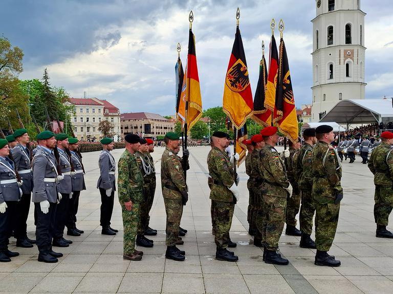 Deutsche Soldaten stehen in Uniformen auf einem öffentlichen Platz in Litauen, einige halten die deutsche Flagge in der Hand. Im Hintergrund ist der Glockenturm der Kathedrale St. Stanislaus in Vilnius zu sehen.