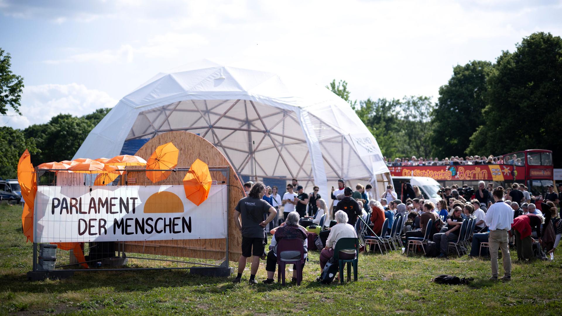 Aktivisten auf der Kundgebung des Parlaments der Menschen in einem kuppelförmigen Zelt. Ein Schild trägt die Aufschrift "Parlament der Menschen" und ist mit orangefarbenen Schirmen bestückt. Aktivisten auf der Kundgebung des Parlaments der Menschen in einem kuppelförmigen Zelt. Ein Schild trägt die Aufschrift "Parlament der Menschen" und ist mit orangefarbenen Schirmen bestückt.