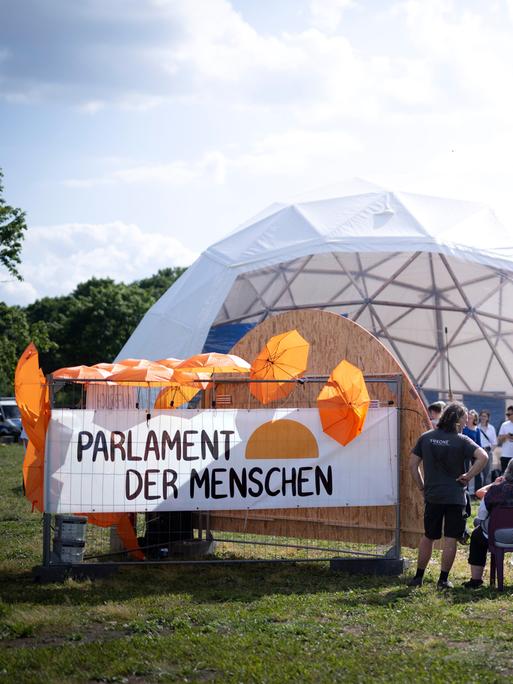Aktivisten auf der Kundgebung des Parlaments der Menschen in einem kuppelförmigen Zelt. Ein Schild trägt die Aufschrift "Parlament der Menschen" und ist mit orangefarbenen Schirmen bestückt.