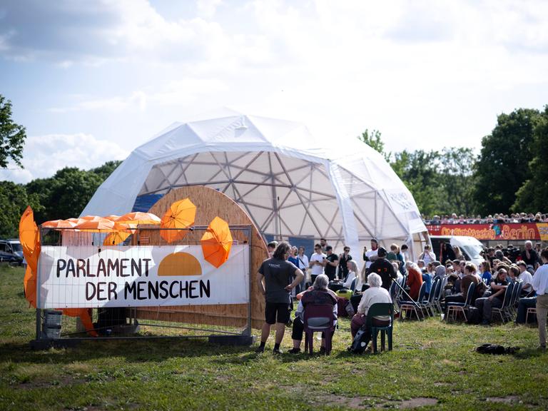 Aktivisten auf der Kundgebung des Parlaments der Menschen in einem kuppelförmigen Zelt. Ein Schild trägt die Aufschrift "Parlament der Menschen" und ist mit orangefarbenen Schirmen bestückt.