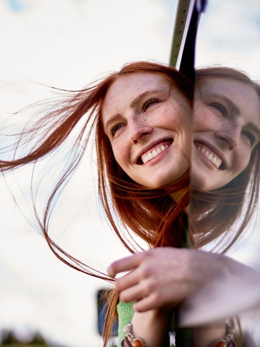 Eine Frau mit langem rotem Haar schaut lachend aus einem offenen Autofenster.