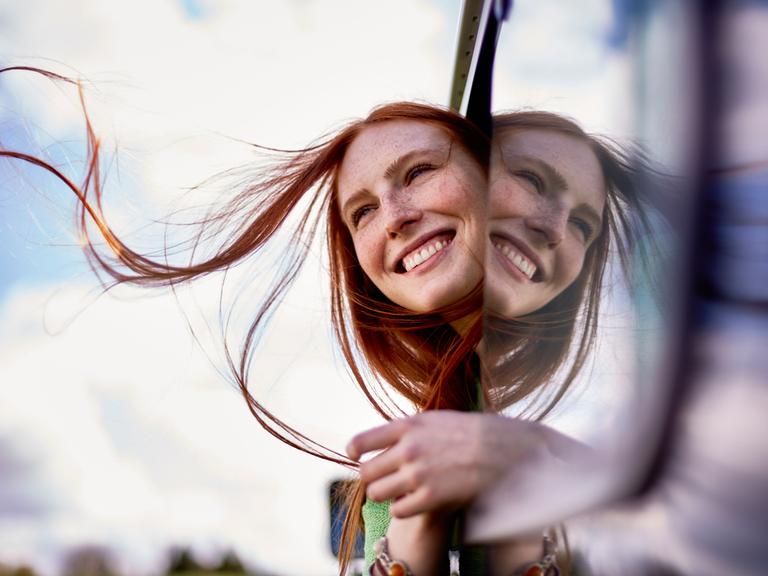 Eine Frau mit langem rotem Haar schaut lachend aus einem offenen Autofenster.