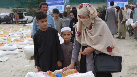 Afghaninnen und Afghanen bekommen kostenlose Nahrungsmittel in Kabul im August 2022. Im Vordergrund eine verschleierte Frau, zwei Jungs und ein Mann, die Getreidesäcke und Speiseöl auf einem Schubkarren wegbringen. Im Hintergrund zahlreiche Menschen, Informationsschilder und aufgestapelte Nahrungsmittel.