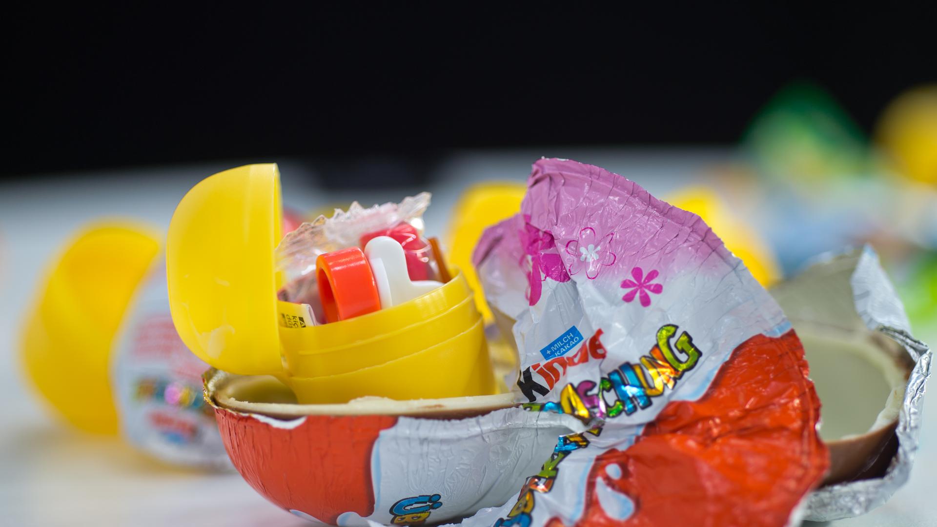 Überraschungseier-Kapseln mit Spielzeug liegen auf einem Tisch zwischen Schokolade und der Verpackung von Kinder-Überraschung. 