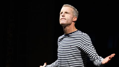 Benjamin von Stuckrad-Barre steht im schwar-weiß gestreiften Pullover mit ausgebreiteten Armen vor einem dunklen Bühnenhintergrund.