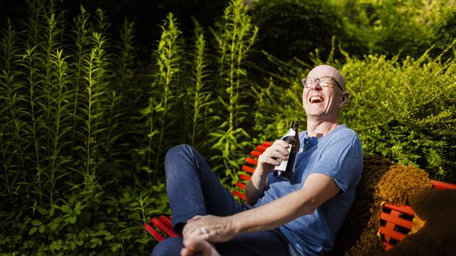 Ein Mann sitzt auf einer Bank im Garten. Er hält eine Bier-Flasche in der Hand und lacht.