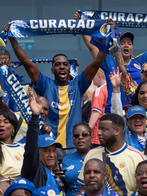 Fans aus Curaçao bei einem Länderspiel zwischen Schottland und Curaçao im Mai 2026. Die Fans feiern fröhlich und halten blaue Curaçaoschals und Fächer.
