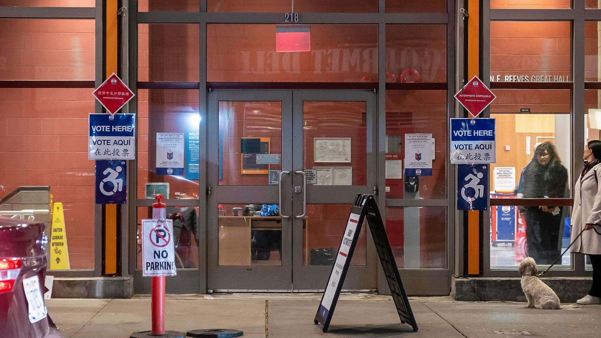 Blick auf ein Wahllokal mit der Aufschrift "Vote Here". Es ist dunkel, die Fenster sind erleuchtet. Vor dem Wahllokal sitzt ein Hund. Blick auf ein Wahllokal mit der Aufschrift "Vote Here". Es ist dunkel, die Fenster sind erleuchtet. Vor dem Wahllokal sitzt ein Hund.